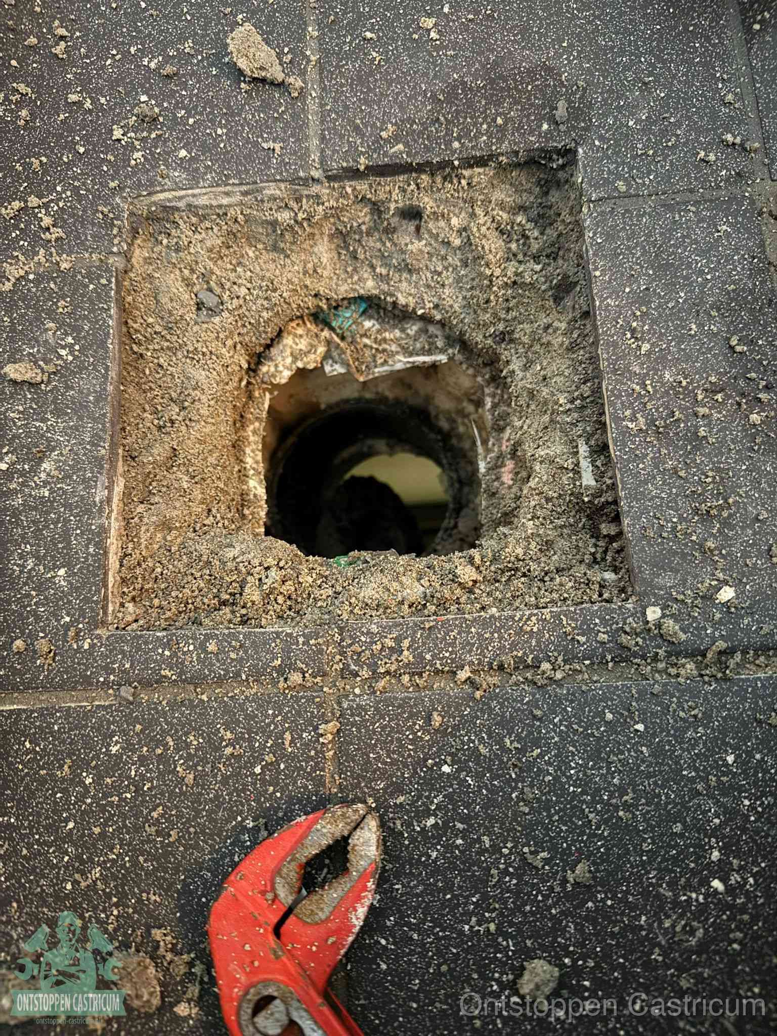 Een geopend rioolputje in een betontegelvloer met losgemaakt rooster en een zichtbare ronde afvoerbuis, het achterhalen waar de afvoerverstopping zit in Castricum