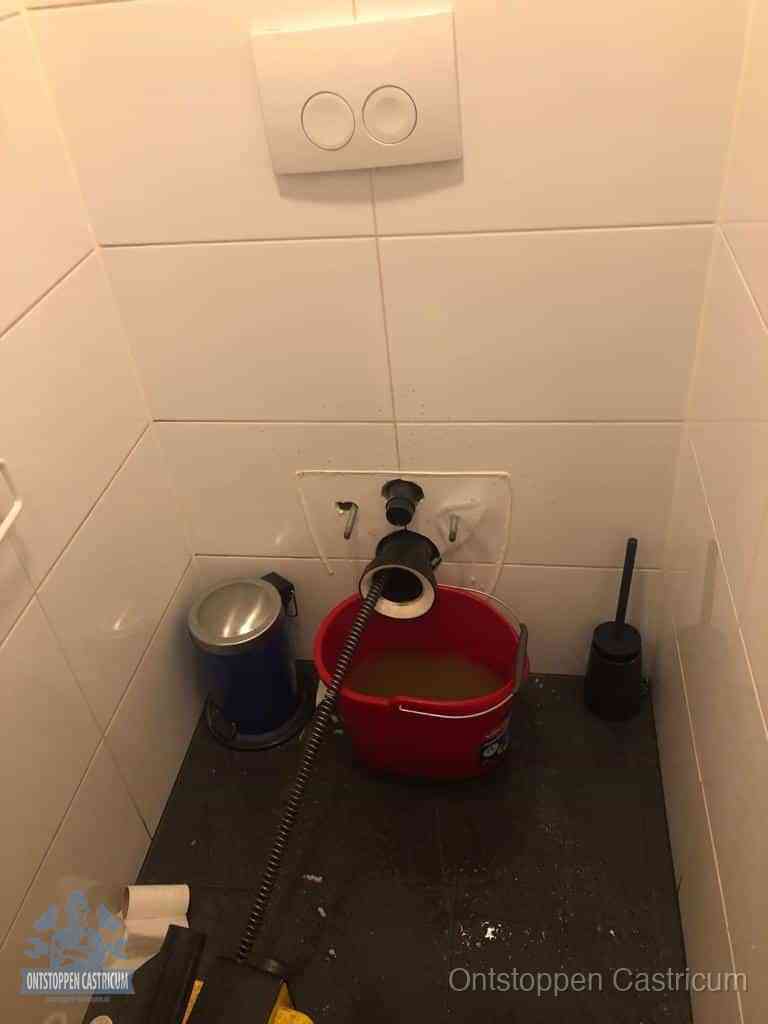 Een ontstopte wand-wc met verwijderde toiletpot waar een ontstoppingsveer in de wanduitgang verdwijnt, het achterhalen waar de verstopping zit in Castricum