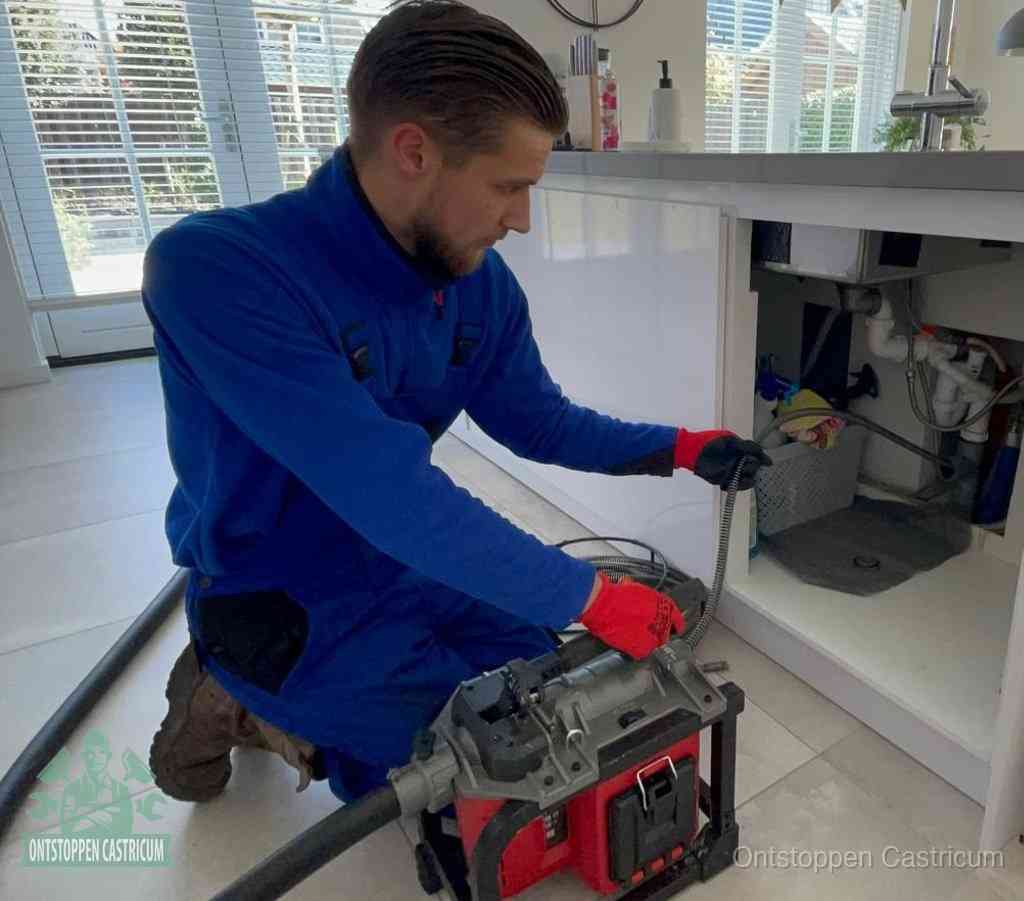 Een monteur met rode handschoenen knielt in een keuken en voert een elektrische ontstoppingsveer in tijdens het professioneel afvoer ontstoppen in Castricum